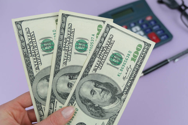 Geld, Amerikaanse bankbiljetten close-up, rekenmachine en bril op paarse achtergrond.. Concept van geld, verdienen, belastingen, twee keer betalen. - Foto, afbeelding