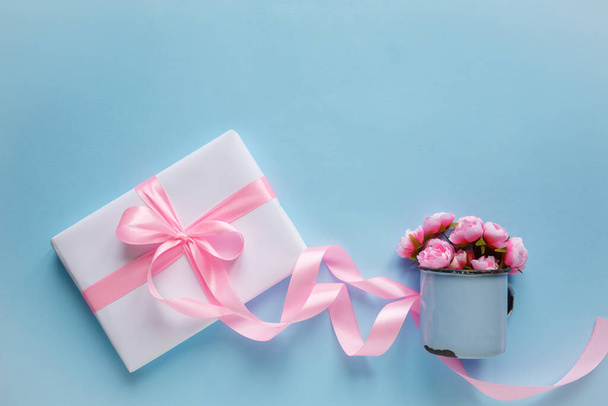 Gift box wrapped in white paper with pink ribbon and vintage blue cup on blue background. Simple composition with presents - 写真・画像