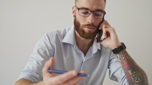 A focused young businessman wearing eyeglasses is talking on the phone during working with paper documents in the white office - Imágenes, Vídeo