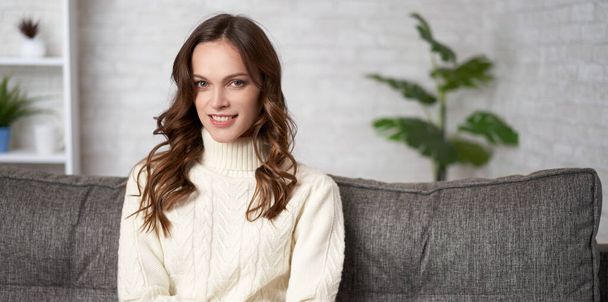 Hermosa mujer sonriente con la piel limpia, maquillaje natural y pelo largo ondulado está usando suéter de punto sentado en un sofá
 - Foto, imagen