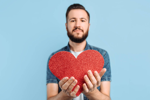 Love and Valentine's day an attractive man with a beard holds a red paper heart, smiling, on an isolated blue background. Valentine's day. Red heart. - Photo, image