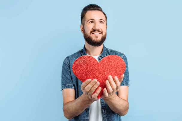 Love and Valentine's day an attractive man with a beard holds a red paper heart, smiling, on an isolated blue background. Valentine's day. Red heart. - 写真・画像