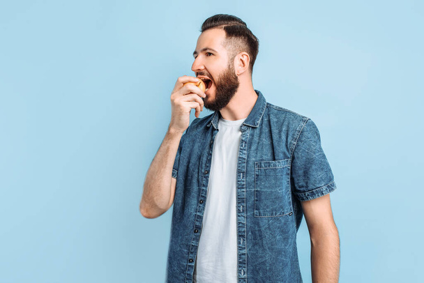 An attractive smiling man bites an Apple and looks at the camera, standing on a blue background - Photo, Image
