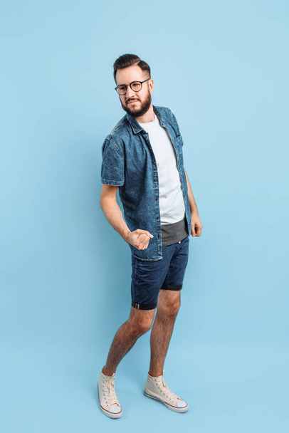 An attractive smart man in glasses, in casual clothes shows a gesture of victory and success, standing on an isolated blue background - 写真・画像