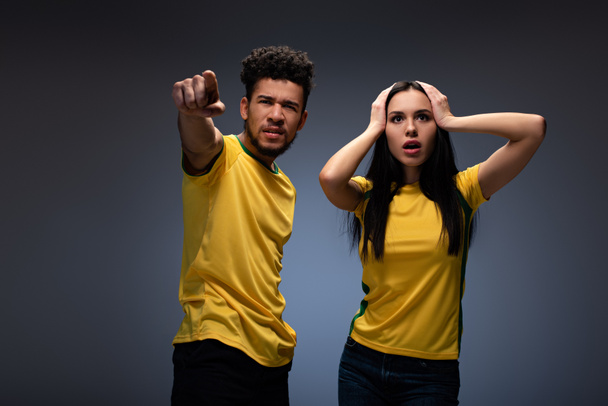 Casal multiétnico preocupado de fãs de futebol em camisetas amarelas apontando em cinza
 - Foto, Imagem