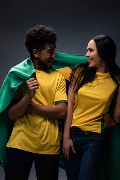 couple of multiethnic smiling football fans with brazil flag on grey - Фото, зображення