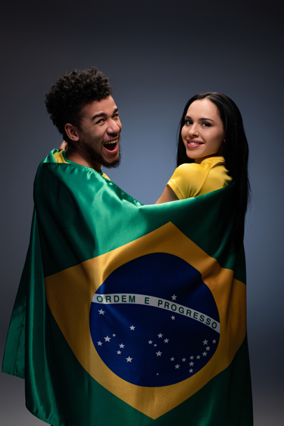 Multikulturelles Paar lächelnder Fußballfans mit brasilianischer Flagge auf grau - Foto, Bild