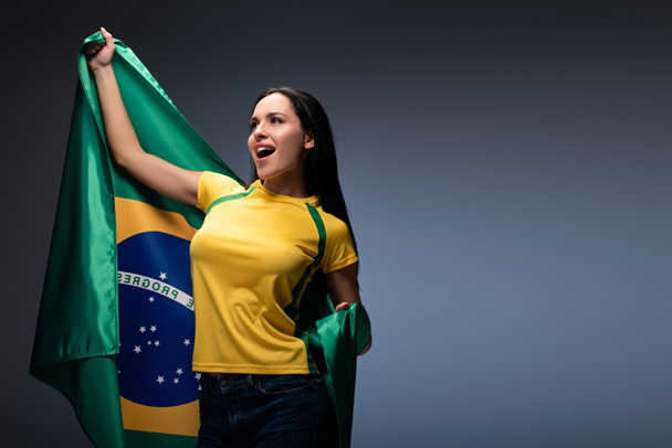 ενθουσιασμένοι θηλυκό ποδοσφαιριστής κρατώντας βραζιλιάνικη σημαία σε γκρι  - Φωτογραφία, εικόνα