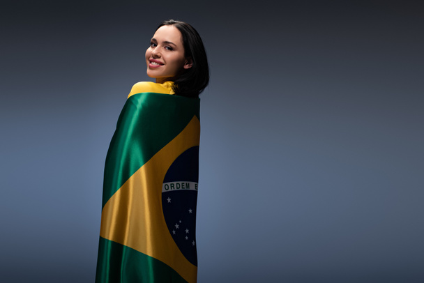 beautiful cheerful female football fan wrapped in brazilian flag on grey - 写真・画像