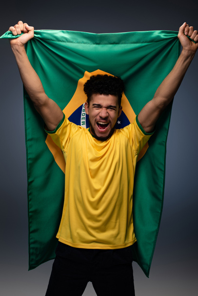opgewonden afrikaanse amerikaanse voetbal fan met Braziliaanse vlag op grijs - Foto, afbeelding