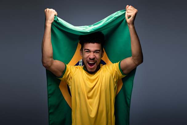 συναισθηματικός Αφροαμερικάνος ποδοσφαιρόφιλος με βραζιλιάνικη σημαία στο γκρι - Φωτογραφία, εικόνα