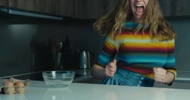A young woman is jumping around and dancing with a spatula in her hand in the kitchen - Záběry, video