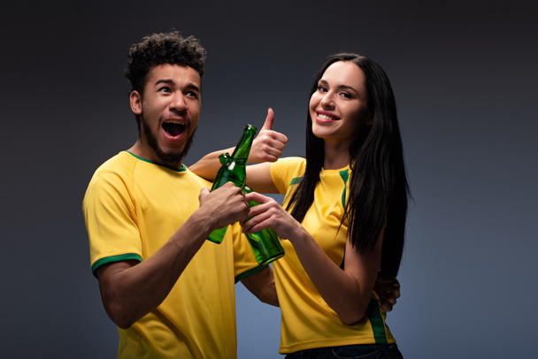 podekscytowana wielonarodowa para kibiców piłki nożnej w żółtych koszulkach brzęczy butelkami piwa i pokazuje kciuk na szarości - Zdjęcie, obraz