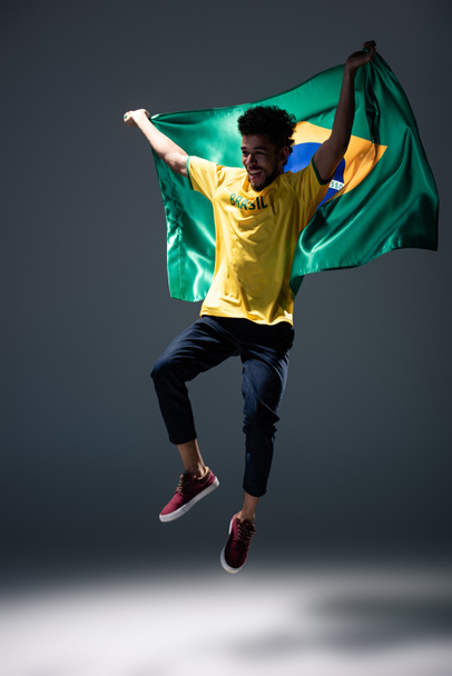 ενθουσιασμένοι Αφροαμερικανοί οπαδοί του ποδοσφαίρου άλμα με βραζιλιάνικη σημαία σε γκρι - Φωτογραφία, εικόνα