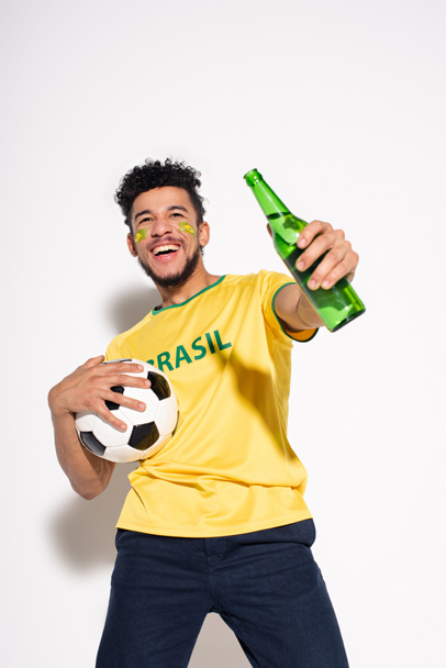 smiling african american man holding football ball and bottle of beer on grey - 写真・画像