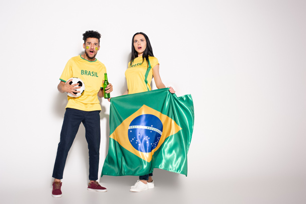 pareja multiétnica de aficionados al fútbol sorprendidos sosteniendo bandera brasileña, bola y botella de cerveza en gris
 - Foto, Imagen