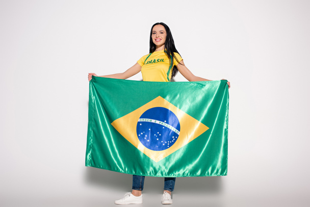 ελκυστική χαμογελαστή γυναίκα οπαδός του ποδοσφαίρου κρατώντας βραζιλιάνικη σημαία σε γκρι - Φωτογραφία, εικόνα