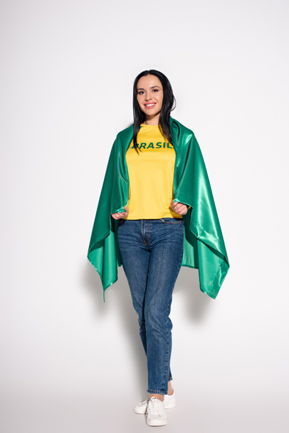 χαμογελαστή γυναίκα ποδοσφαιρόφιλη τυλιγμένη με βραζιλιάνικη σημαία στο γκρι - Φωτογραφία, εικόνα