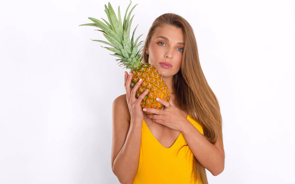 Portret seksownej, długowłosej dziewczyny w żółtym stroju kąpielowym, która pozuje z ananasem w dłoniach na białym tle - Zdjęcie, obraz