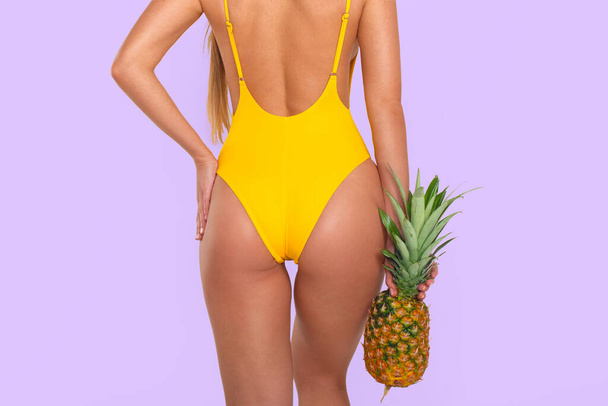Gewassen foto van sexy gebruind meisje in geel badpak poseren met haar rug naar de camera met ananas in haar handen op witte achtergrond  - Foto, afbeelding