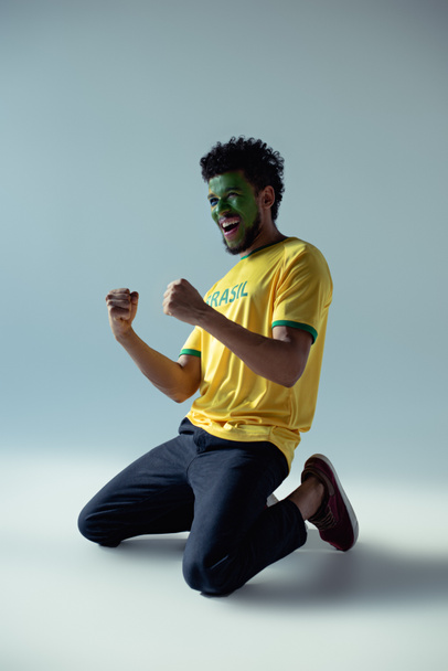 emocionální africký americký fotbalový fanoušek s obličejem namalovaným jako brazilská vlajka klečí na šedé - Fotografie, Obrázek