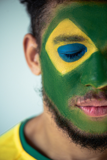 knap Afrikaans amerikaans voetbal fan met gesloten ogen en gezicht geschilderd als Braziliaanse vlag geïsoleerd op grijs - Foto, afbeelding