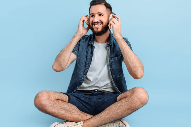 Happy young handsome man in casual clothes listening to music with headphones, sitting on a blue background - Photo, Image