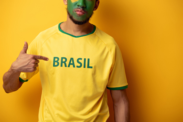 cropped view of football fan with painted face pointing at t-shirt with brazil sign on yellow - Foto, immagini