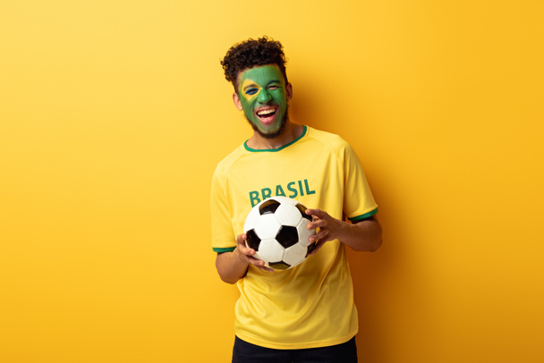 excited african american football fan with face painted as brazilian flag holding ball on yellow - Φωτογραφία, εικόνα