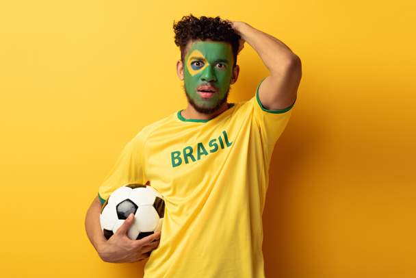 geschokt Afro-Amerikaanse voetbalfan met gezicht geschilderd als Braziliaanse vlag met bal op geel - Foto, afbeelding