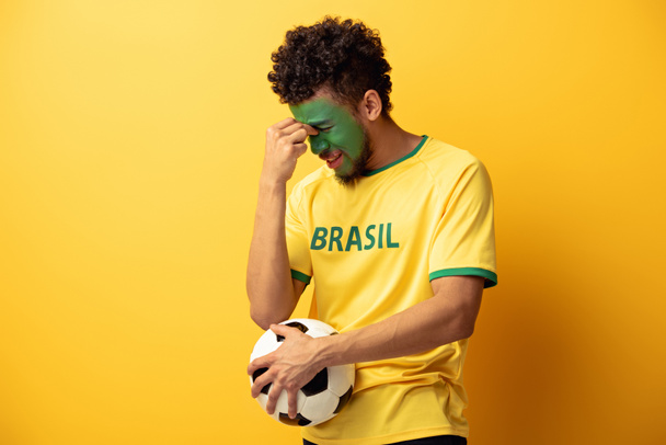 confused african american football fan with face painted as brazilian flag holding ball on yellow - Zdjęcie, obraz