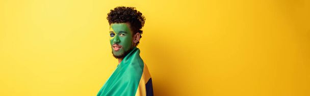 panoramic shot of african american football fan with painted face wrapped in brazilian flag on yellow - Photo, Image