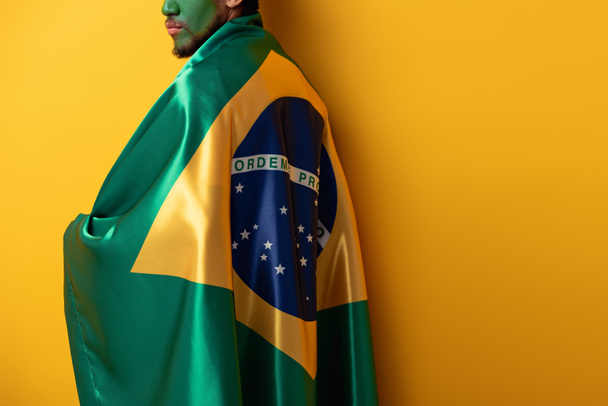 περικοπή άποψη του ανεμιστήρα ποδοσφαίρου με ζωγραφισμένο πρόσωπο τυλιγμένο σε βραζιλιάνικη σημαία σε κίτρινο - Φωτογραφία, εικόνα