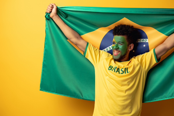 happy african american football fan with painted face holding brazilian flag on yellow - Photo, Image