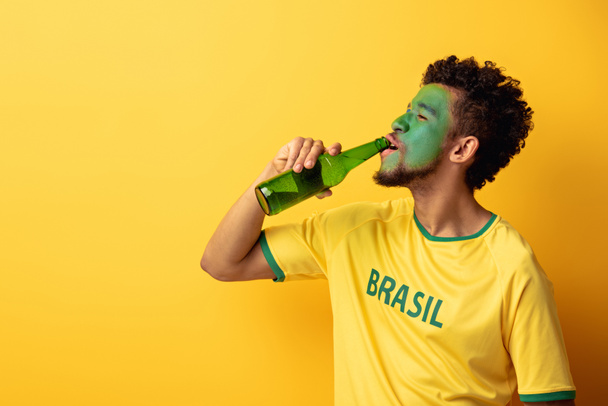heureux fan de football afro-américain avec le visage peint comme drapeau brésilien boire de la bière sur jaune
 - Photo, image