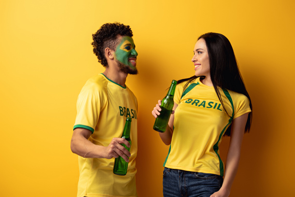 man afrikaanse amerikaanse voetbal fan met geschilderd gezicht en glimlachende vrouw met flessen bier op geel  - Foto, afbeelding