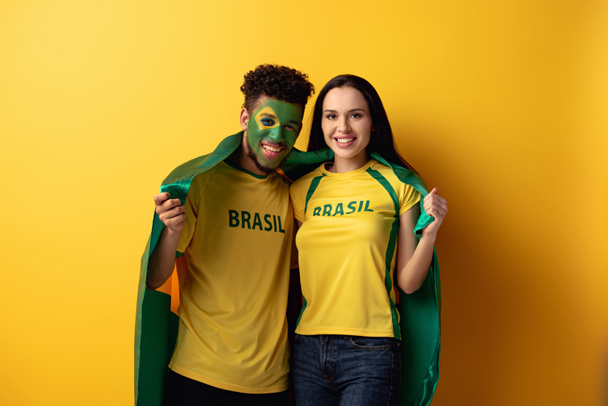 male african american football fan with painted face and smiling girl holding brazilian flag on yellow  - Foto, Bild