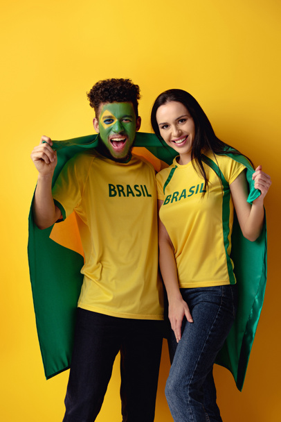schöner männlicher afrikanisch-amerikanischer Fußballfan mit gemaltem Gesicht und glücklichem Mädchen mit brasilianischer Flagge auf gelb  - Foto, Bild