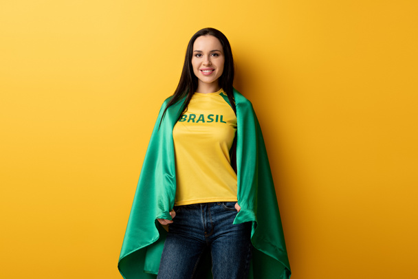 smiling female football fan wrapped in brazilian flag on yellow - 写真・画像