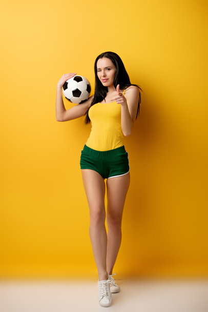 όμορφη γυναίκα οπαδός του ποδοσφαίρου με μπάλα κλείσιμο του ματιού και δείχνοντας σας στο κίτρινο - Φωτογραφία, εικόνα
