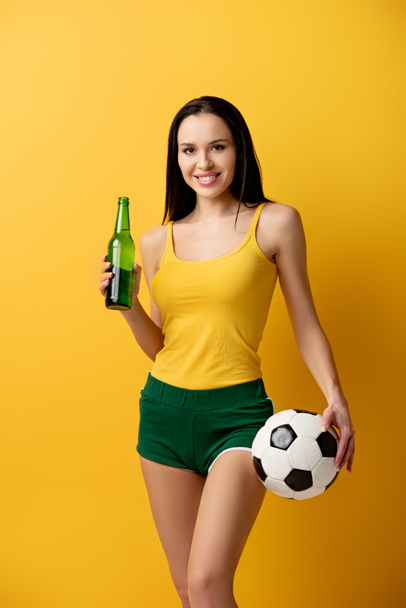 ventilador de fútbol femenino sonriente sosteniendo bola y botella de cerveza en amarillo
 - Foto, imagen