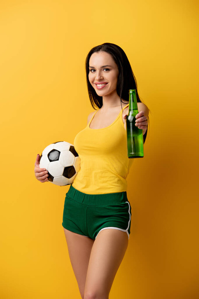 smiling football fan holding ball and bottle of beer on yellow - Fotó, kép