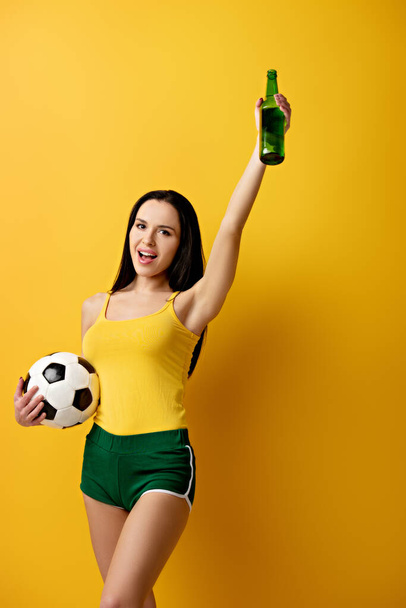 opgewonden vrouwelijke voetbal fan houden bal en fles bier op geel - Foto, afbeelding