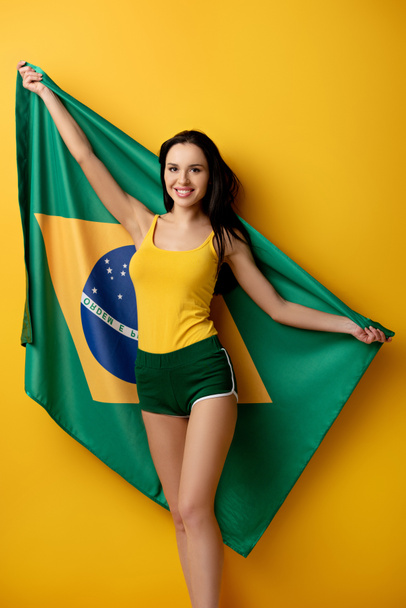 Şortlu neşeli bayan futbol fanatiği sarıda Brezilya bayrağı taşıyor.  - Fotoğraf, Görsel