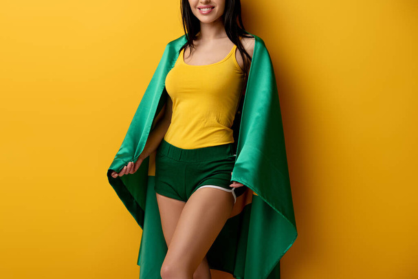 cropped view of smiling female football fan in shorts holding brazilian flag on yellow  - Fotografie, Obrázek