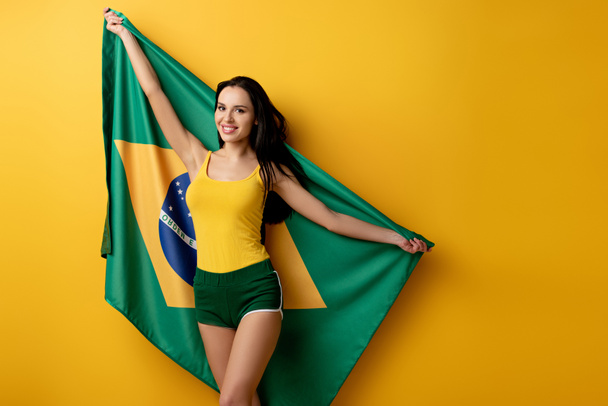smiling female football fan in shorts holding brazilian flag on yellow  - Photo, Image