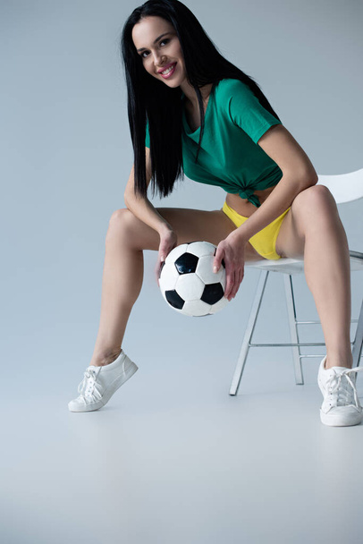 σέξι χαμογελαστή γυναίκα κρατώντας μπάλα ποδοσφαίρου, ενώ κάθεται στην καρέκλα σε γκρι - Φωτογραφία, εικόνα