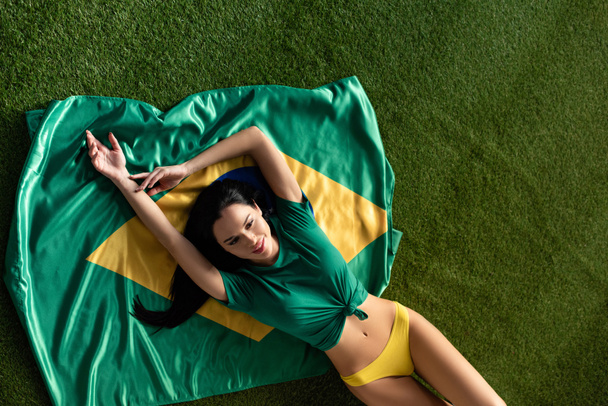 widok góry szczęśliwy sexy dziewczyna leżące na brazylijskiej flagi na zielonej trawie  - Zdjęcie, obraz