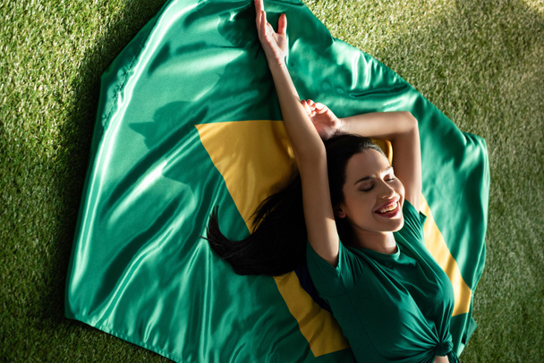χαμογελαστό σέξι κορίτσι ξαπλωμένο στη βραζιλιάνικη σημαία στο πράσινο γρασίδι  - Φωτογραφία, εικόνα