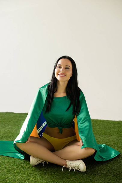 happy sexy football fan sitting with brazilian flag on green grass isolated on grey - Photo, Image